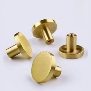 May include: Four brass cabinet knobs with a round, flat face and a cylindrical stem. The knobs are a warm gold color and are arranged against a white background. These knobs are suitable for drawers and cabinets.