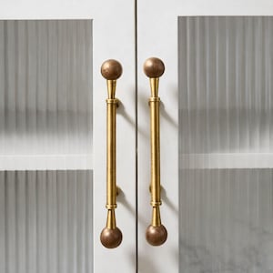 May include: Close-up of a white cabinet with two doors, each featuring a long, gold-colored handle. The handles have a cylindrical design with a spherical knob at each end. The cabinet doors have glass panels with vertical ribbed detailing.