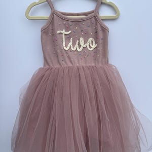 May include: A dusty rose-colored toddler dress with a tulle skirt and spaghetti straps. The bodice has the word "two" embroidered in white, with colorful confetti-like accents. The dress is hanging on a wooden hanger.
