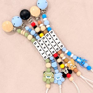 May include: Five pacifier clips featuring round wooden or plastic clips in light blue, black, and wood grain. Each clip has a string of colorful silicone beads, white letter beads spelling DAVID, and a character charm including Stitch, Simba, Mickey, and cars.