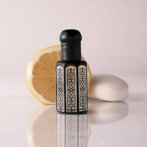 May include: A black and cream-colored perfume bottle with an ornate design stands in front of a lemon slice and a white bar of soap. The bottle has a black cap and a decorative pattern.