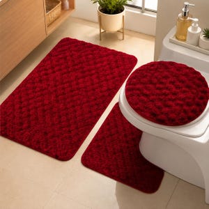May include: A three-piece bathroom rug set in a deep red color, featuring a rectangular rug, a U-shaped contour rug, and a round toilet lid cover. Each piece has a textured, tufted surface with a consistent pattern throughout the entire set.