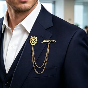 May include: A gold-toned metal lapel pin featuring a lion head emblem and a nameplate with the text Antonio, connected by two draped chains. The accessory is attached to the lapel of a dark navy blue suit jacket worn over a white dress shirt.