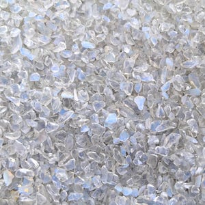 May include: Close-up of a pile of small, irregularly shaped, clear glass pieces with a subtle, iridescent sheen. The pieces are scattered, creating a textured surface with hints of blue and white.
