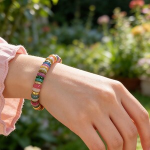 May include: A colorful beaded bracelet with a variety of gemstones, including red, green, and blue stones, is worn on a wrist. The bracelet has gold-colored spacers. The background is a blurred garden scene with green foliage and flowers.