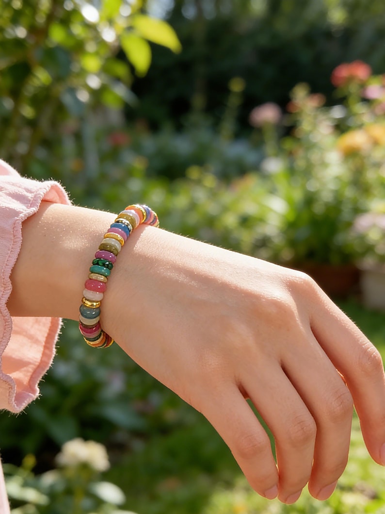 May include: A colorful beaded bracelet with a variety of gemstones, including red, green, and blue stones, is worn on a wrist. The bracelet has gold-colored spacers. The background is a blurred garden scene with green foliage and flowers.