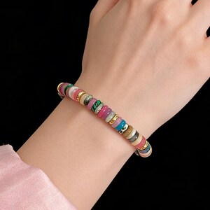 May include: A colorful beaded bracelet with a variety of round, flat beads in shades of pink, green, blue, and gold. The bracelet is worn on a wrist against a black background. The beads are approximately 0.25 inches in diameter.