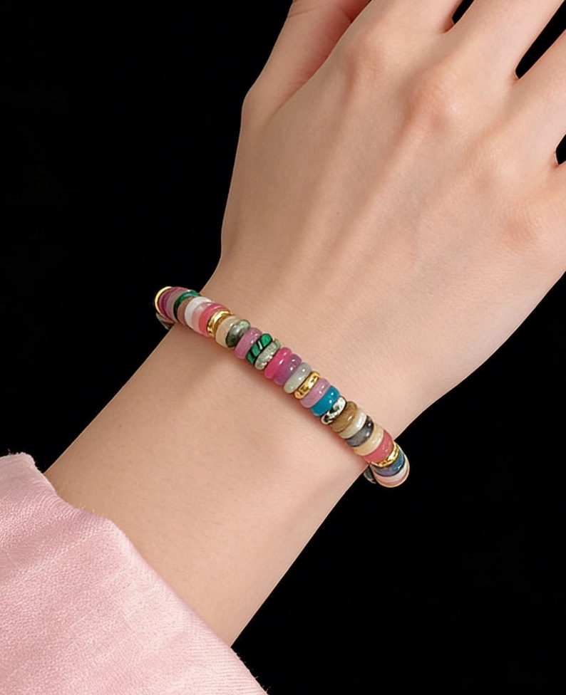 May include: A colorful beaded bracelet with a variety of round, flat beads in shades of pink, green, blue, and gold. The bracelet is worn on a wrist against a black background. The beads are approximately 0.25 inches in diameter.