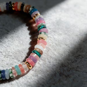 May include: A colorful beaded bracelet featuring a variety of gemstones in shades of pink, green, blue, and gold. The bracelet is made of small, round beads strung together with gold-colored spacers. The bracelet is shown on a soft, white surface.