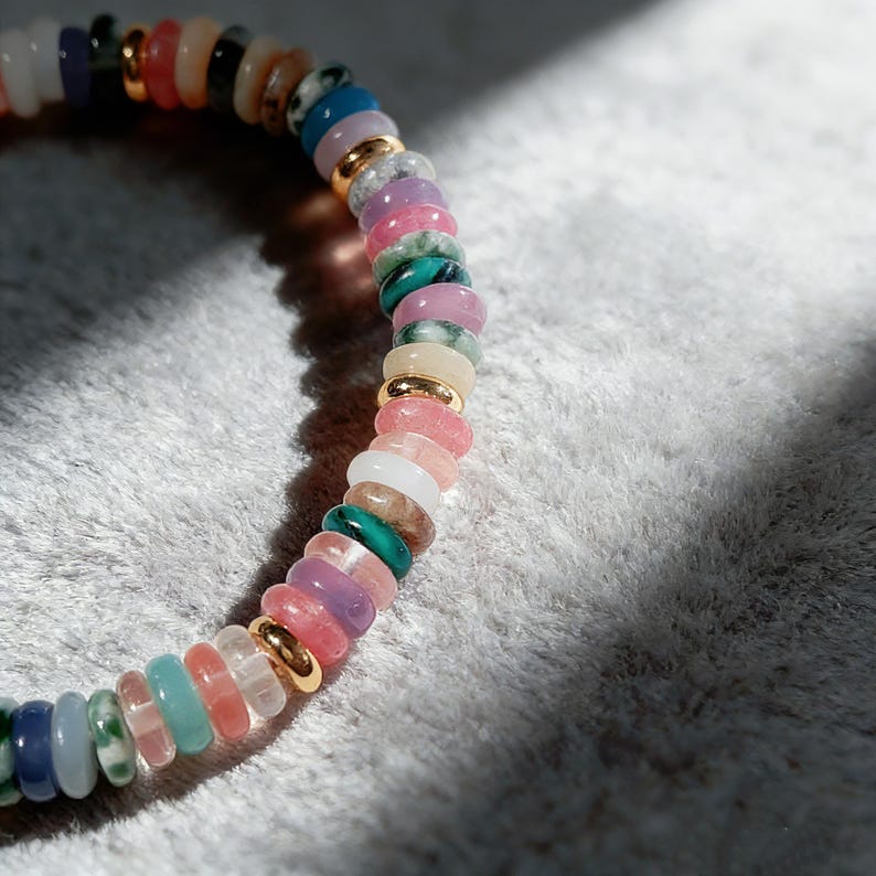 May include: A colorful beaded bracelet featuring a variety of gemstones in shades of pink, green, blue, and gold. The bracelet is made of small, round beads strung together with gold-colored spacers. The bracelet is shown on a soft, white surface.