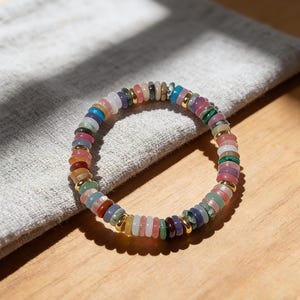 May include: A colorful beaded bracelet with a variety of gemstones. The bracelet features a mix of round, flat beads in shades of pink, green, blue, purple, and yellow, separated by gold-colored spacers. The bracelet is resting on a gray cloth.