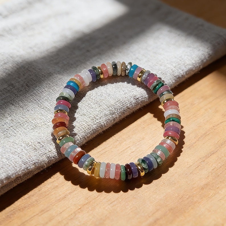 May include: A colorful beaded bracelet with a variety of gemstones. The bracelet features a mix of round, flat beads in shades of pink, green, blue, purple, and yellow, separated by gold-colored spacers. The bracelet is resting on a gray cloth.