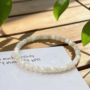 May include: A delicate bracelet composed of small, round, off-white beads. The bracelet rests on a white card with partial text visible. The background is a light-colored wooden surface with green leaves in the upper corners.