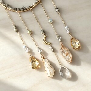 May include: A gold-toned wire hoop adorned with gray stone chips, featuring five hanging chains. The chains include small gold stars, a crescent moon, clear and gray beads, two wire-wrapped raw crystals, and three faceted amber and clear teardrop pendants.