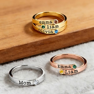 May include: Three personalized rings in gold, rose gold, and silver. The gold ring has three bands with the names "emma", "liam", and "olivia" and colored gemstones. The rose gold ring has "Emma" and "Liam" with gemstones. The silver ring has "Mom" and a clear gemstone.