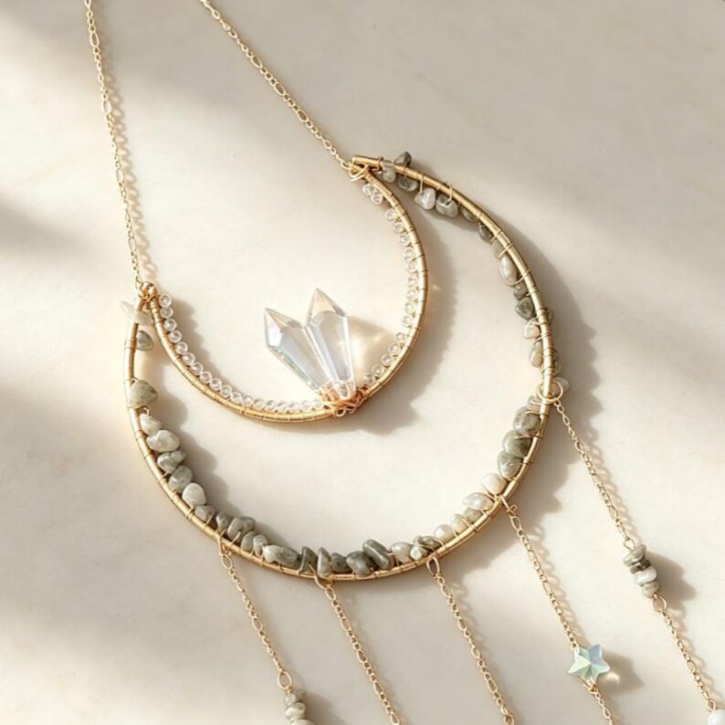 May include: A gold-toned necklace featuring two crescent-shaped wire frames adorned with small grey stone chips and clear beads. The inner crescent holds two clear crystal points, while the outer crescent has dangling gold chains with small star charms.