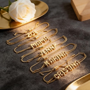 May include: Gold-colored name bracelets spelling out "ZOE", "MAMA", "JENNY", "KARINA", and "CRISTINA". The bracelets are on a gray surface, with a gold tray and a white rose in the background. The bracelets have adjustable chains.
