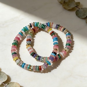 May include: Two beaded bracelets with a variety of colorful gemstones. The bracelets feature a mix of pink, green, blue, yellow, and purple beads, separated by small gold-colored spacers. The bracelets are displayed on a white marble surface.