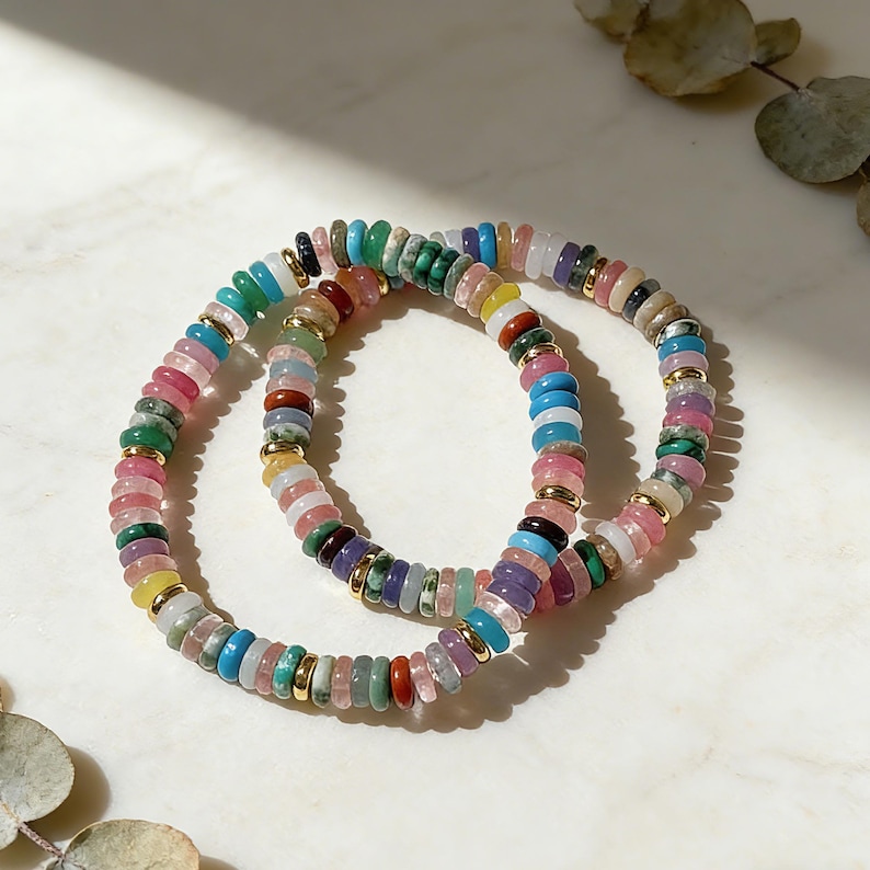 May include: Two beaded bracelets with a variety of colorful gemstones. The bracelets feature a mix of pink, green, blue, yellow, and purple beads, separated by small gold-colored spacers. The bracelets are displayed on a white marble surface.