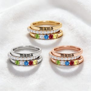 May include: Three stacked rings in gold, silver, and rose gold. Each ring features the word "mama" and a row of colorful gemstones. The rings are set against a white, textured background.
