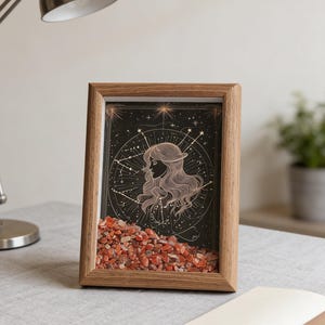 May include: A framed art piece featuring a woman's profile within a celestial design. The artwork is set against a dark background with gold accents and a layer of orange-red stones at the base. The frame is made of light-colored wood.