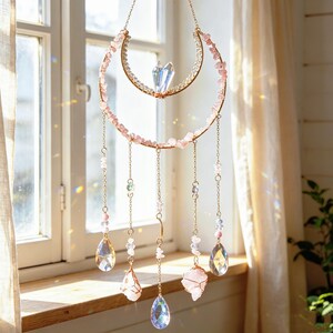 May include: A hanging suncatcher featuring a gold-toned crescent moon frame adorned with small pink stone chips. Five delicate gold chains dangle from the frame, each ending in either a clear teardrop-shaped crystal or a raw pink stone pendant.