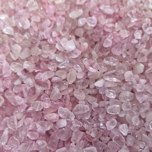 May include: A close-up of a collection of small, polished rose quartz stones. The stones are a soft pink colour and vary in size and shape. The surface is filled with the stones, creating a textured appearance. The stones have a translucent quality.
