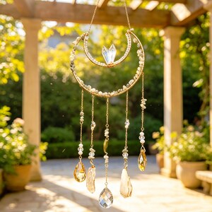 May include: A crescent moon-shaped wall hanging featuring a gold-toned wire frame adorned with small light-colored stones and a central clear crystal point. Five delicate gold chains hang from the frame, each decorated with beads and clear glass teardrop prisms.