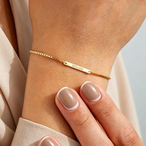 May include: A gold-toned chain bracelet featuring a thin, rectangular metal bar centred on the chain. The bar is engraved with the name WILLIAM in a clean, sans-serif font. The bracelet has a delicate, adjustable box chain design.