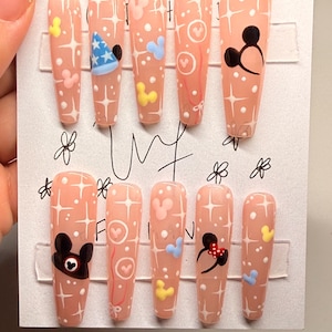 May include: Set of ten artificial nails with a light peach base, adorned with white starbursts, Mickey Mouse and Minnie Mouse designs, and other cartoon elements. The nails are arranged on a white card.