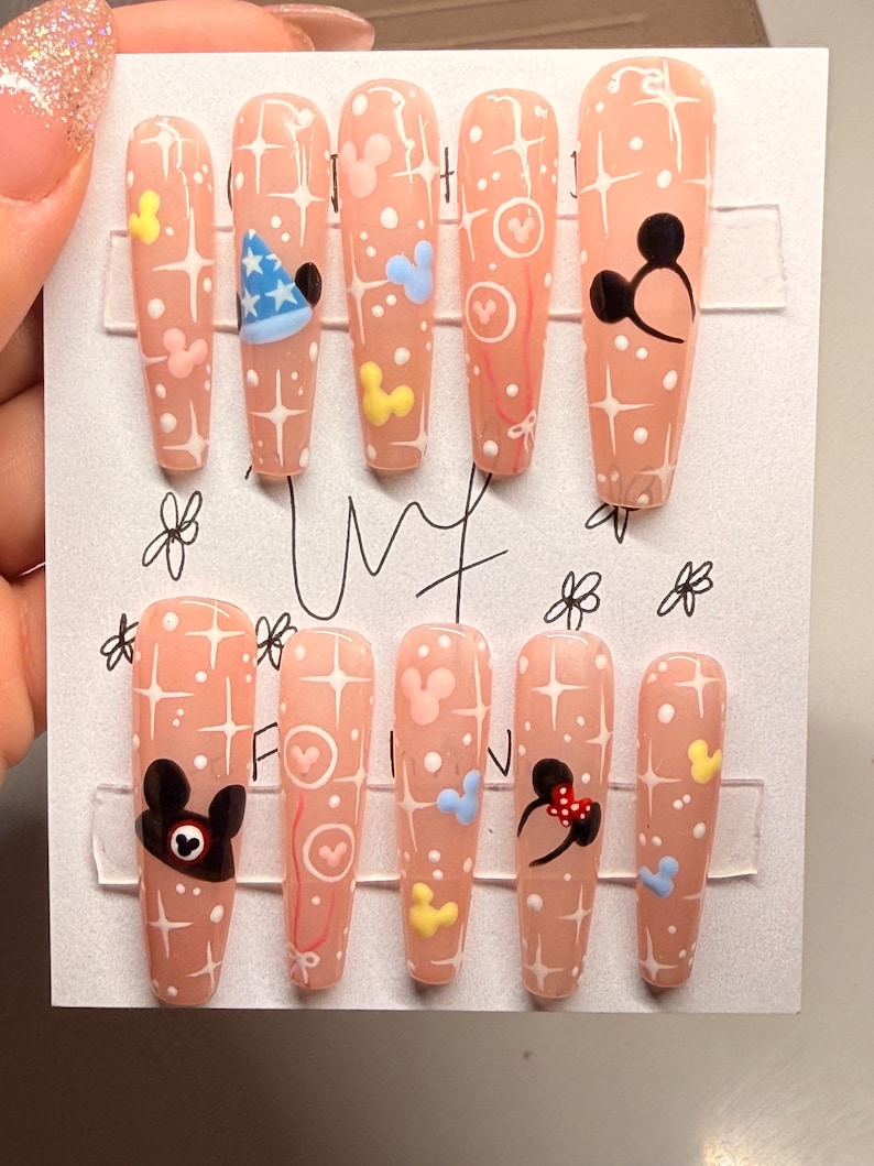 May include: Set of ten artificial nails with a light peach base, adorned with white starbursts, Mickey Mouse and Minnie Mouse designs, and other cartoon elements. The nails are arranged on a white card.