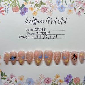 May include: A set of almond-shaped, short, press-on nails with a light pink base. Each nail features a different design, including hearts, Mickey Mouse, and Stitch. The text "Wildflower Nail Art" is at the top.