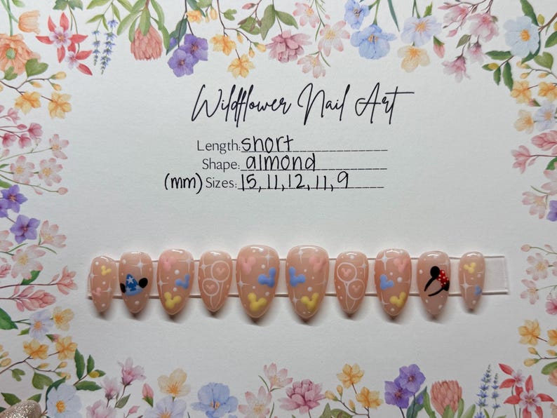 May include: A set of almond-shaped, short, press-on nails with a light pink base. Each nail features a different design, including hearts, Mickey Mouse, and Stitch. The text "Wildflower Nail Art" is at the top.