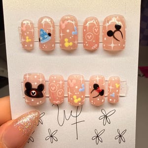 May include: A set of press-on nails with a light pink base, featuring cartoon character designs. Designs include a blue party hat, hearts, and Mickey Mouse-inspired graphics. The nails are arranged on a white card.