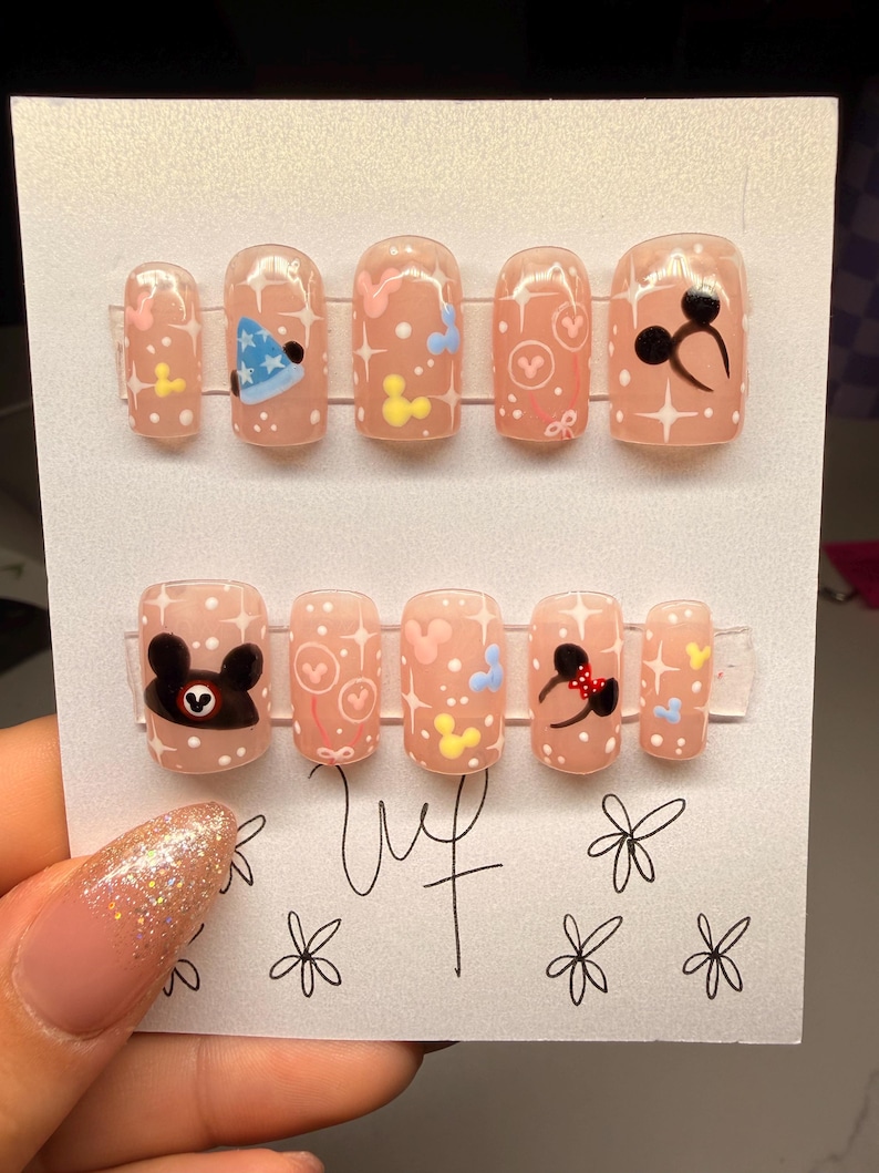 May include: A set of press-on nails with a light pink base, featuring cartoon character designs. Designs include a blue party hat, hearts, and Mickey Mouse-inspired graphics. The nails are arranged on a white card.