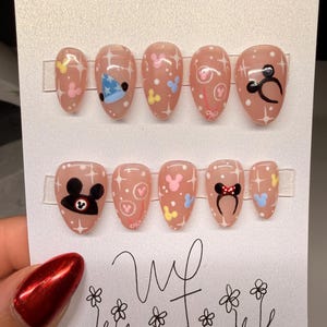 May include: A set of ten oval-shaped, press-on nails in a light pink hue. Each nail features a different design, including Mickey Mouse and Minnie Mouse-themed graphics, stars, and polka dots. The nails are displayed on a white card.