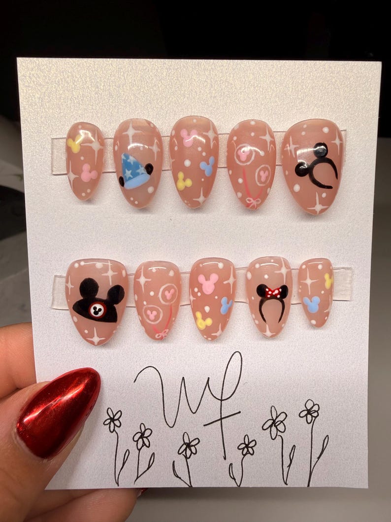 May include: A set of ten oval-shaped, press-on nails in a light pink hue. Each nail features a different design, including Mickey Mouse and Minnie Mouse-themed graphics, stars, and polka dots. The nails are displayed on a white card.
