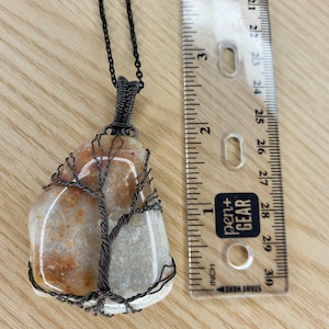 May include: A handcrafted pendant necklace featuring a polished, irregularly shaped stone wrapped in dark wire. The wire is shaped into a tree design. The pendant is approximately 2 inches long, suspended from a dark chain.