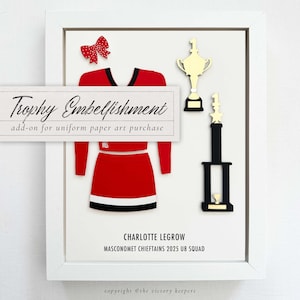 Puede incluir: Obra de arte en papel enmarcada con un uniforme de animadora rojo y negro, un lazo rojo y dos trofeos dorados. El texto dice "Trophy Embellishment" y "Charlotte Legrow, Masconomet Chieftains 2025 U8 Squad."