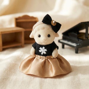 Puede incluir: Una figura de oveja con una parte superior negra con un diseño de copo de nieve blanco y una falda dorada. La oveja tiene un lazo negro con un detalle dorado y una diadema beige. Muebles en miniatura y un piano en el fondo.
