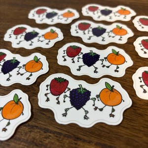 May include: A collection of whimsical stickers featuring a strawberry, a blackberry, and an orange, each with cartoon legs, arranged in a row. The stickers are white with a scalloped edge, and the background is a wooden surface.