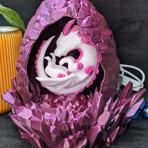 Peut inclure: Une sculpture fantastique d'un dragon éclosant d'un œuf. L'œuf et la base sont d'un violet profond avec des formations cristallines. Le dragon est blanc avec des accents roses.