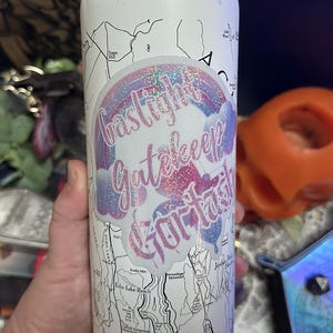 Peut inclure: Une gourde blanche avec un motif de carte et un autocollant avec le texte "Gaslight Gatekeep Gorl Task" dans une police arc-en-ciel. La gourde est cylindrique et semble être en métal. L'arrière-plan comprend un crâne orange et d'autres objets.