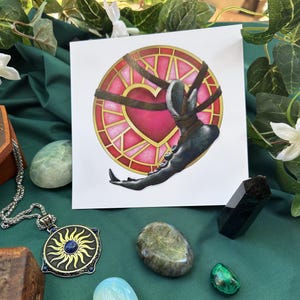 May include: A square art print featuring a heart design within a stained glass-style circle, with a dark figure wrapped around it. The image is surrounded by crystals, a pendant necklace, and greenery on a green cloth.