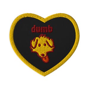 May include: Heart-shaped embroidered patch with a black centre and a gold border. The word "dumb" is stitched in red above a yellow dog illustration with red accents. Suitable for clothing or accessories.