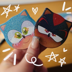 May include: Two cartoon character stickers, one blue and one black, held by a hand. The blue character has green eyes and a smiling face. The black character has red eyes and a stern expression. White star and heart doodles surround the stickers.