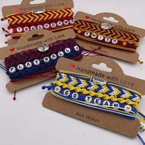 May include: Four handmade friendship bracelets with sports-themed words. The bracelets are red, blue, yellow, and orange, with white letter beads spelling "TOUCHDOWN", "PLAY BALL", "SWISH", and "GO TEAM". Each bracelet is on a brown card with the text "Handmade with Love" and "Best Wishes".