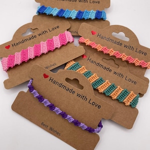 May include: A collection of handmade macrame bracelets in various colors, including blue, pink, orange, green, and purple. Each bracelet is displayed on a brown card with the text "Handmade with Love" and "Best Wishes".
