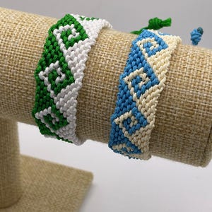 Könnte beinhalten: Zwei handgefertigte Freundschaftsarmbänder. Eines ist grün und weiß, das andere blau und cremefarben. Beide Armbänder weisen ein gewebtes Muster auf. Die Armbänder werden auf einer beigefarbenen, strukturierten Oberfläche präsentiert.