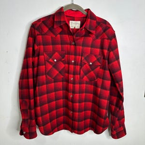 May include: A long-sleeved button-down shirt featuring a red and black buffalo plaid pattern. The shirt includes a pointed collar, two chest pockets with buttoned flaps, and silver-toned metal snap buttons down the front and on the cuffs.